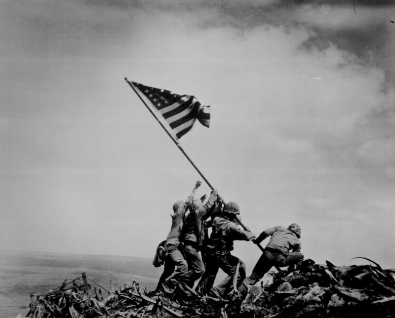 Marines raise an American flag on a rocky, rubble-strewn island.