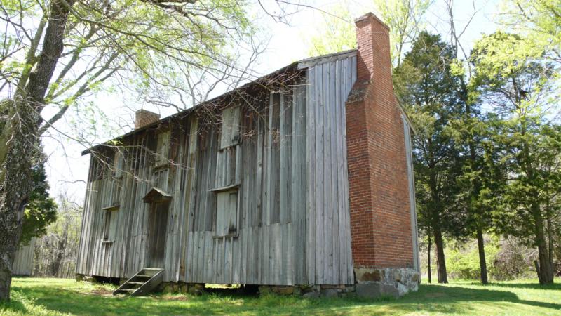 A ramshackle wooden shack with a meager chimney sits against a woodland. It is sunny.