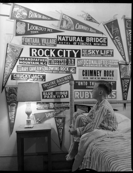 Photograph of Hugh Morton's son, Jim. He is sitting on his bed looking at the tourism stickers collected on his wall.