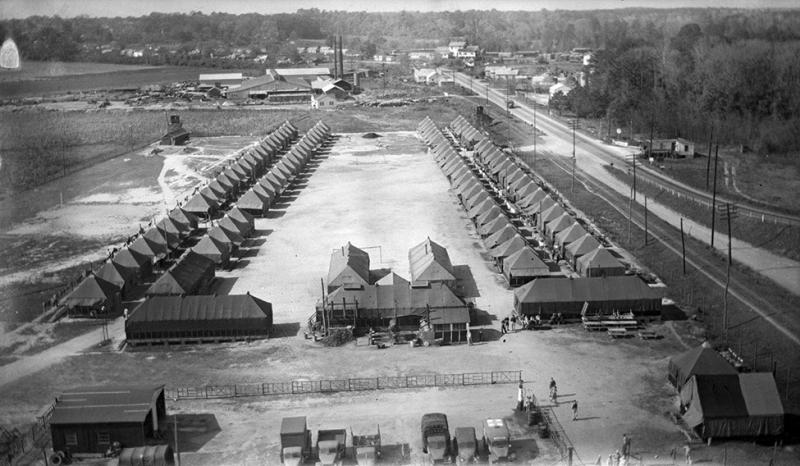 A prisoner of war camp. It is in the middle of a field. There are about 40 barracks on each side with some buildings in the front. There are trees around and some automobiles parked in front of the buildings in the camp. Black and white. 