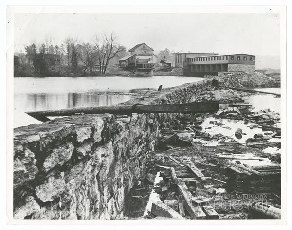 Idol dam. A reservoir of water is held by a stone wall. A facility sits at the top of the reservoir next to a home. Trees surround the reservoir. 