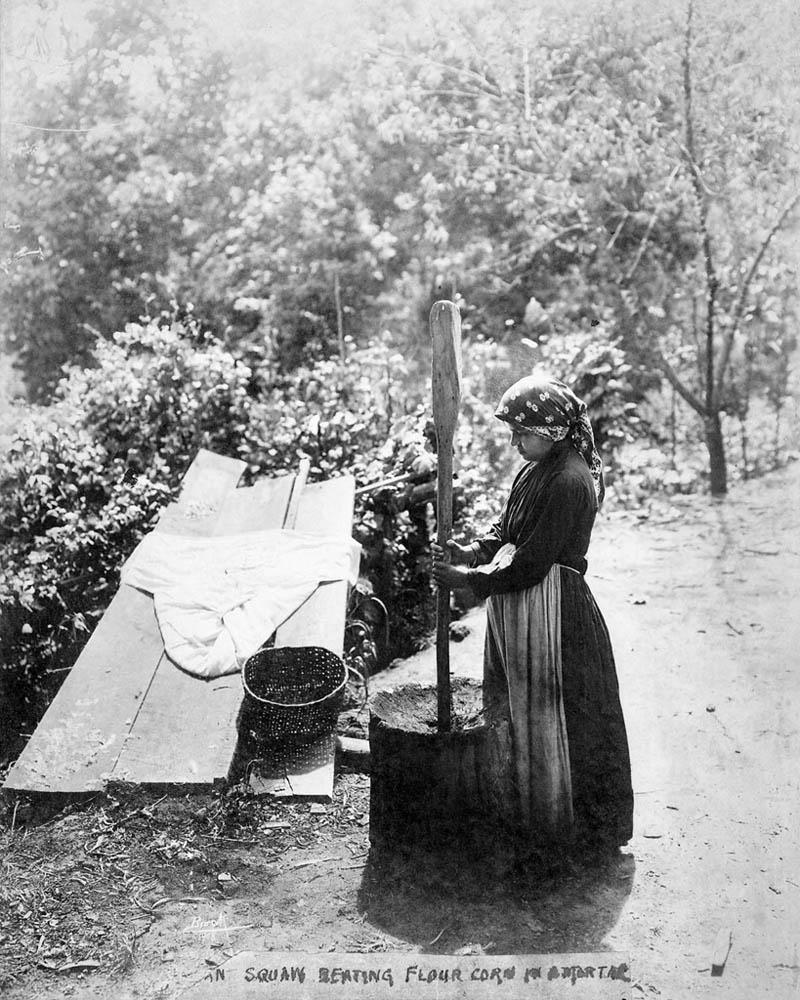 Black and white photograph of an American Indian woman pounding corn with a large mortar and pestle.