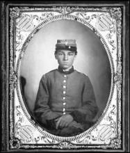 Portrait of a Confederate soldier. He is young, and wearing a grey jacket and hat. His hands are crossed and he looks concerned. He has no facial hair. 