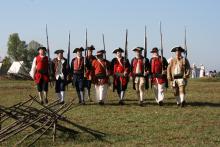 Nine reenactor soldiers in a formation. They are wearing apparel of the colonial military, with tricorn hats and buttoned coats. It is sunny.