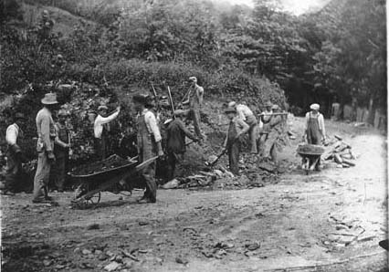 Black and white photograph of men in overalls working to build road. They are using shovels and wheelbarrows. 