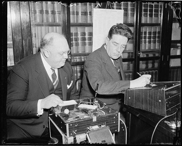 "Demonstrates new signature machine to house member. Washington, D.C., March 18 [1938]. The latest is the automatic electric signature machine invented by Glenn W. Watson, (left) who is shown in this picture demonstrating it to Rep. Lindsay C. Warren of North Carolina." Presented on Library of Congress.
