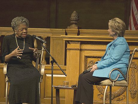 Maya, seated left, speaks into a microphone. She is wearing a black dress and has permed hair. Hillary is seated right, wearing a sky blue blazer and listening intently. 