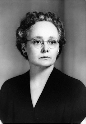 Mary Scott with an updo, glasses, and a dark, V-necked top. Her expression is serious.