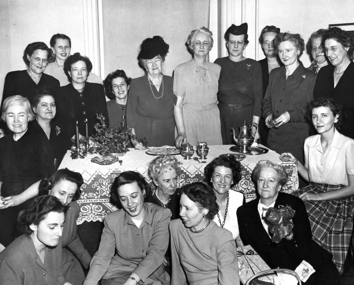 In the black and white photograph a group of women pose around a lace covered dining table. The table is set with a silver tea service, a plate of thin wafers, and a Christmas decoration. The women are all dressed up and some hold Christmas presents. The woman standing in the center back with white hair and glasses and wearing a short-sleeved polka-dotted dress is Jane McKimmon. The women sitting almost directly in front of her but on the other side of the table with dark hair and long chain-link necklace is Ruth Current.