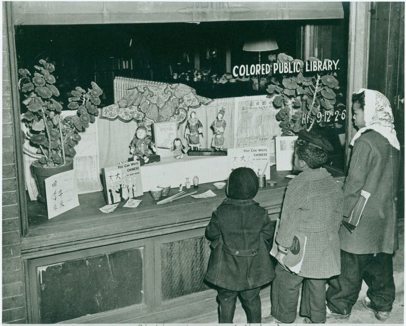 Three young African-American children carrying schoolbooks ("Essentials of Everyday English") stand looking into the display window of the Market Street Branch Library in the YMI building in Asheville. The library branch, first called the "Colored Library," opened in 1927 on South Market Street. The name was changed to Market Street Branch of City Libraries in 1951. The branch moved to another part of the YMI building, facing Eagle Street, in 1959. That branch closed in 1966, following the desegregation of the library system in 1962. Window is stenciled: "Colored Public Library - Hrs 9-12, 2-6." The subject of the window display is China, with plants, dolls, newspapers, and books. The names of the children are (left to right) Spencer White, Frank Owens Jr., and Dorothy Owens. They were students at the Shiloh Elementary School in Asheville at the time this photo was taken.