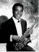 A young black man sits holding a saxophone.  He is wearing a suit coat and white button-up shirt.  He is smiling.