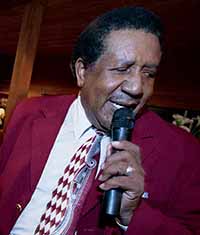 An older black man speaks into a microphone.  He is wearing a dark red suit, white button-up shirt, and a patterned tie.