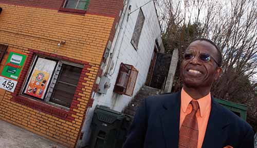 A black man stands next to a brick building.  He is wearing a dark suit coat, peach colored button-up shirt, a dark orange tie, and sunglasses.  He is smiling.