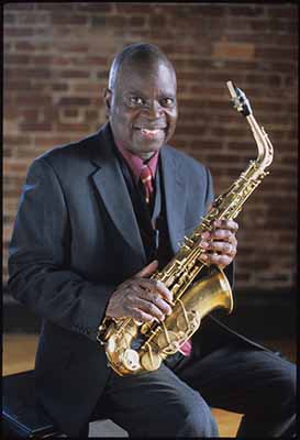 A black man sits holding a saxophone.  He is wearing a dark suit, vest, dark red button-up shirt, and dark red tie.  He is looking at the camera smiling.