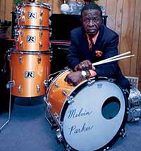 An older black man sits with a drum set.  He is wearing a dark suit, with an orange button-up shirt and a tie.  He is leaning on a a large drum holding drumsticks.  Three smaller drums are stacked next to him.