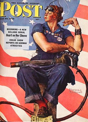 A woman sits in a worksuit with a sandwich in her hand. Her hair is tied and she has welding goggles on her head.
