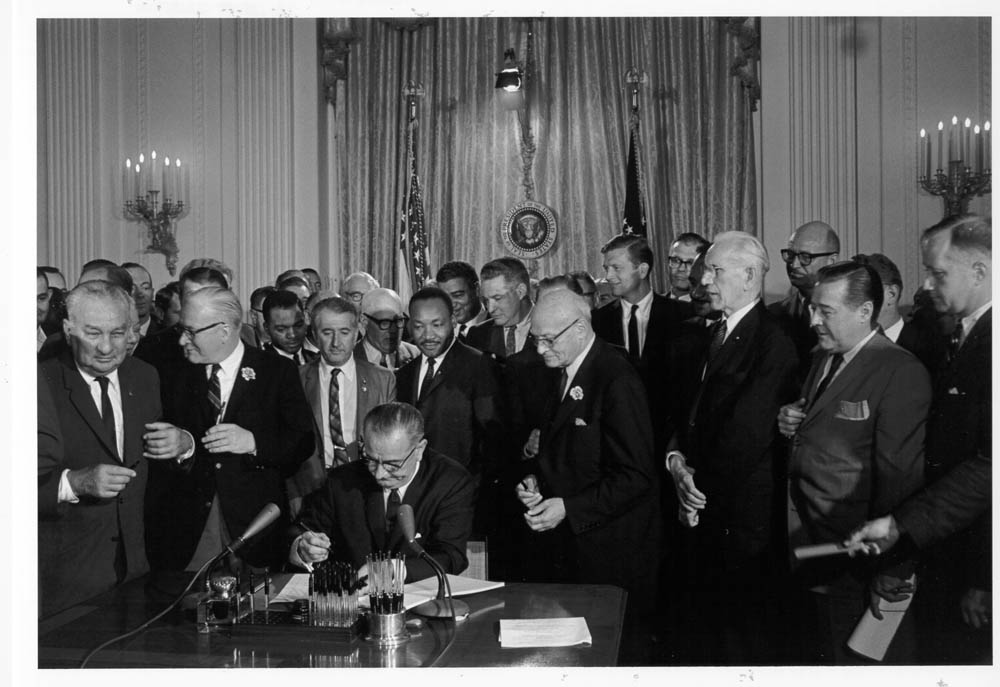 President Lyndon Johnson signs legislation on his desk. He is surrounded by other dignitaries.