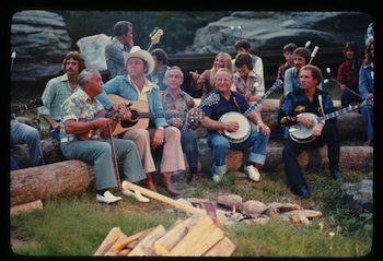 Five men surround a campfire and are playing instruments. There are people and large rocks in the background. 