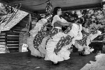 Venezuelan dancers perfoming. Three women on stage with a barn in the background. They have white dresses and are actively dancing. 