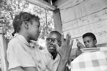 A community worker for the North Carolina Fund teaches math to two children on the front porch of their home near Boone in 1964. Photograph by Billy Barnes. North Carolina Collection, University of North Carolina at Chapel Hill Library.