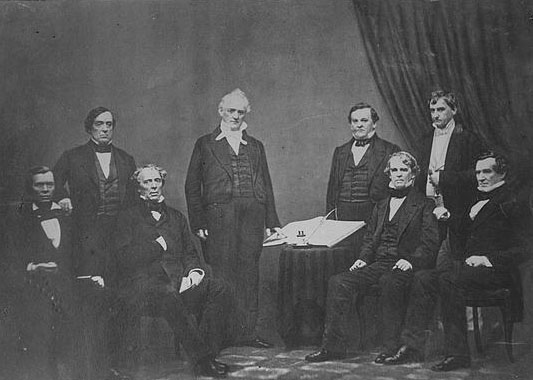"Buchanan, Pres. & cabinet. Photograph shows President Buchanan standing, surrounded by his Cabinet including Jacob Thompson, Secretary of the Interior; Lewis Cass, Secretary of State; Howell Cobb, Secretary of the Treasury; Jeremiah Black, Attorney General; Horatio King, Postmaster General; John B. Floyd, Secretary of War and Isaac Toucey, Secretary of the Navy." Presented on Library of Congress.
