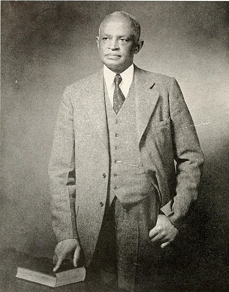 Dr. Shepard. He is standing with his hand on a stack of books while wearing a suit. He looks older.