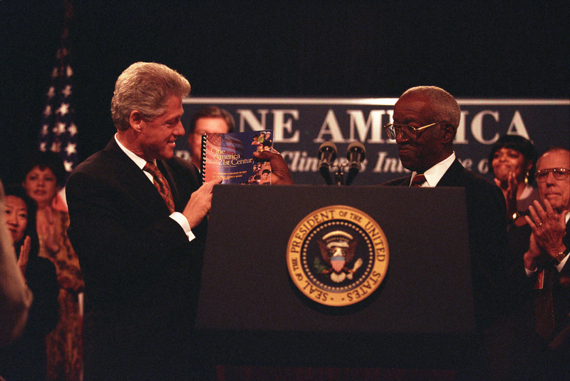 President Clinton stands left holding a book, while John Hope Franklin stands right, holding the same book. They are both wearing suits and are being applauded by onlookers.