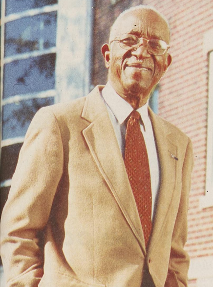 John Hope Franklin wearing a tan suit and smiling. He is older, and a brick building is behind him.