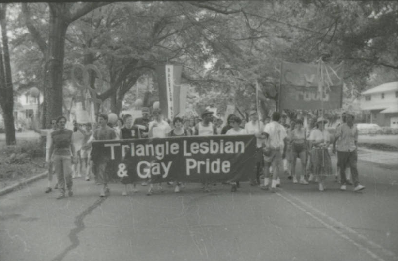 Protestors marching on a street. They hold a banner that says "Triangle Lesbian and Gay Pride."