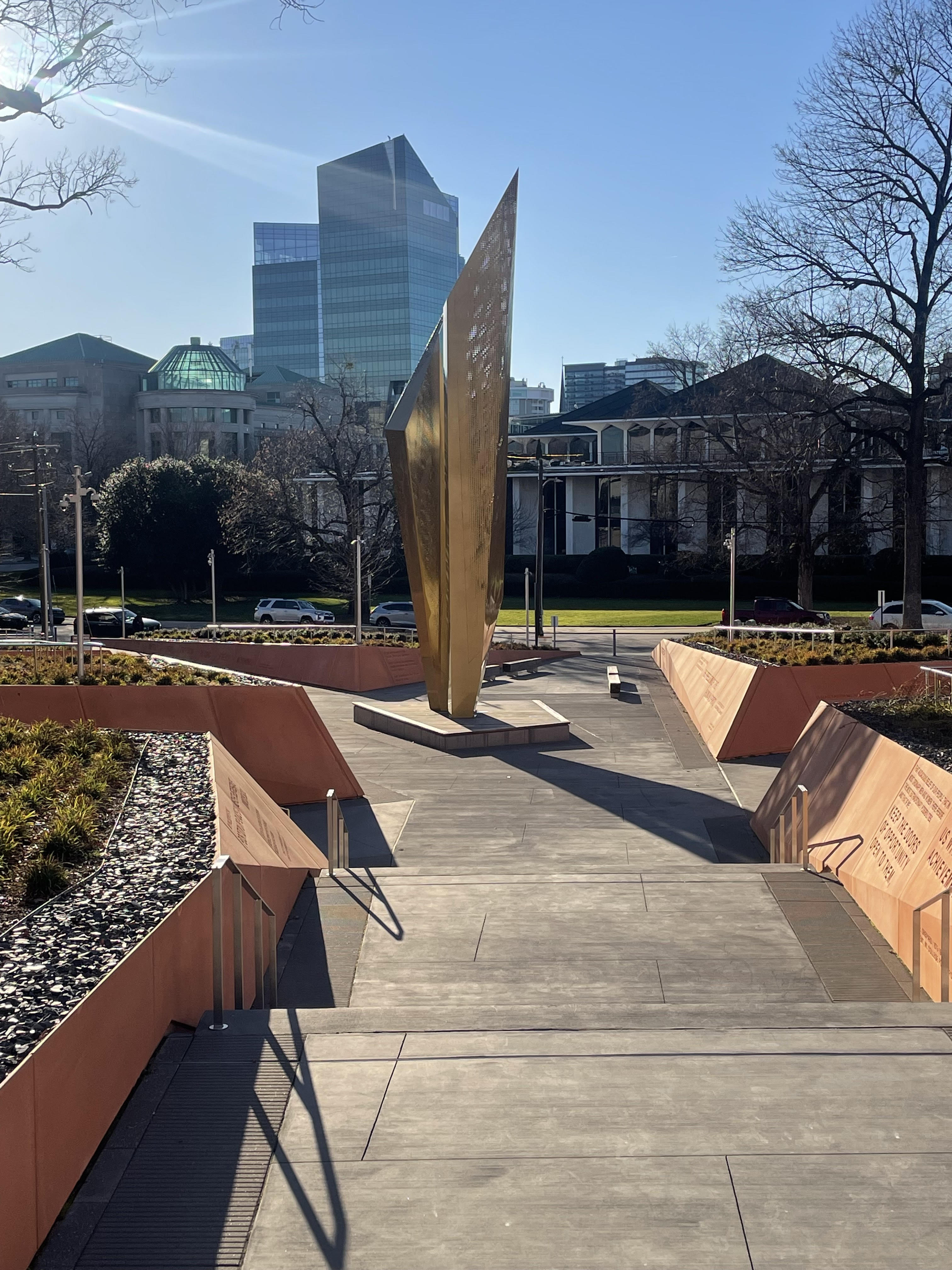 A park where a large golden beacon sits in the middle. Trees and bushes dot the foreground, while a city skyline marks the background..