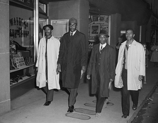 Four men walking. They are the Greensboro Four. They are all sharply dressed with coats and hats. They have determined looks on their faces. 