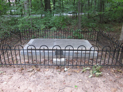 A small stone grave, surrounded by wrought iron fence. Flanked by a woodline.