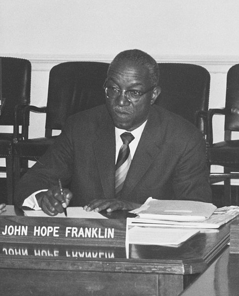 Franklin seated at a desk. A name placard with his name printed on it is in front of him. He is holding a pen and presiding over some papers. He looks stern. 