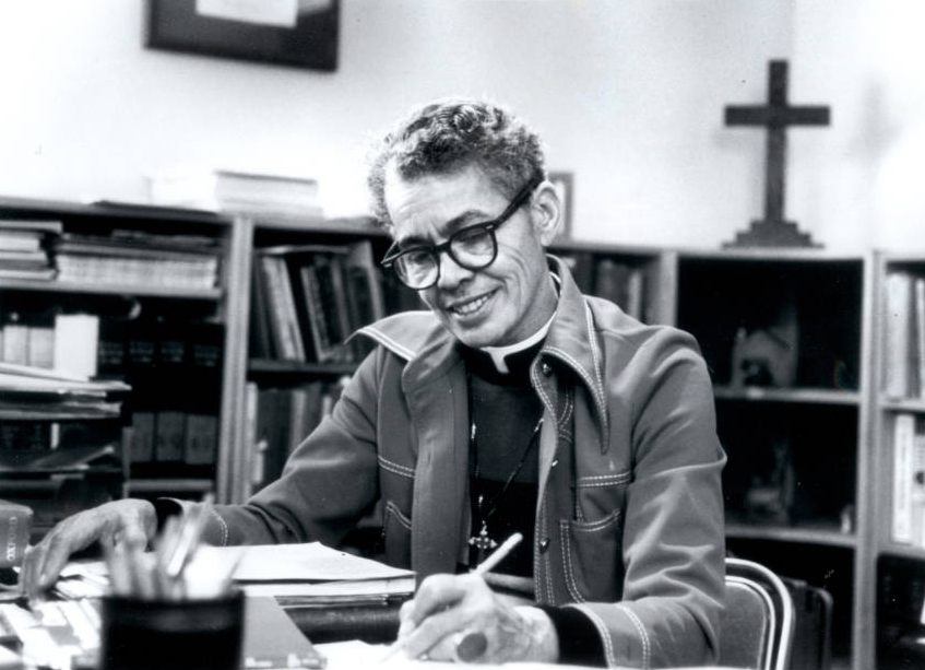 Pauli Murray at a desk, pen in hand. Pauli is smiling, wearing glasses, and writing. Pauli appears to be in an office.