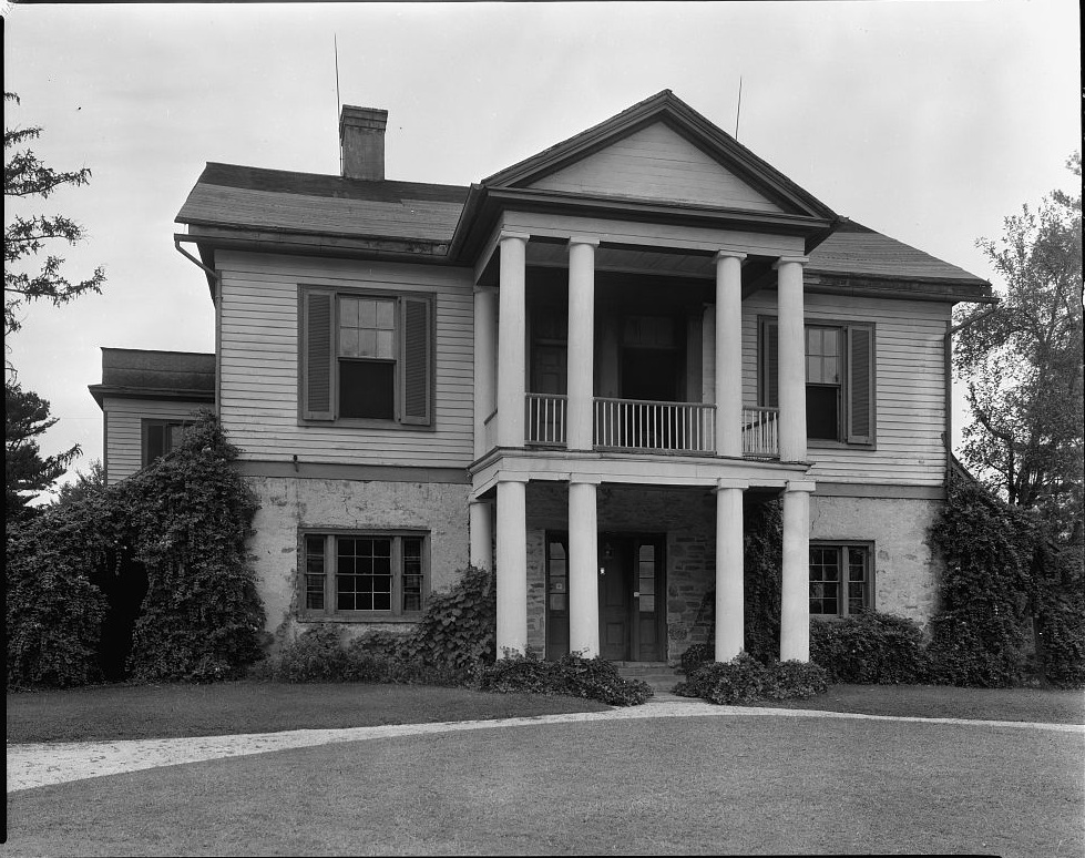 Front view of the Struan house.  A stone path leads up to a columned entryway.  The columns of the first floor support a second floor balcony.  Windows are on either side of the entry and balcony. The first floor is constructed of stone and the second floor of siding. A chimney is visible on the left side of the house.