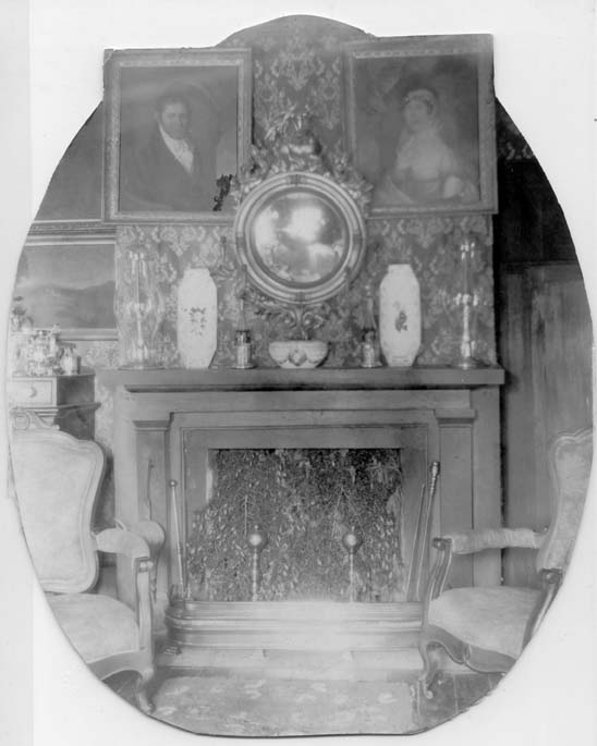 A fireplace with vases on the mantle.  A round mirror and two portraits hang above the mantel. Two upholstered armchairs sit in front of the fireplace.