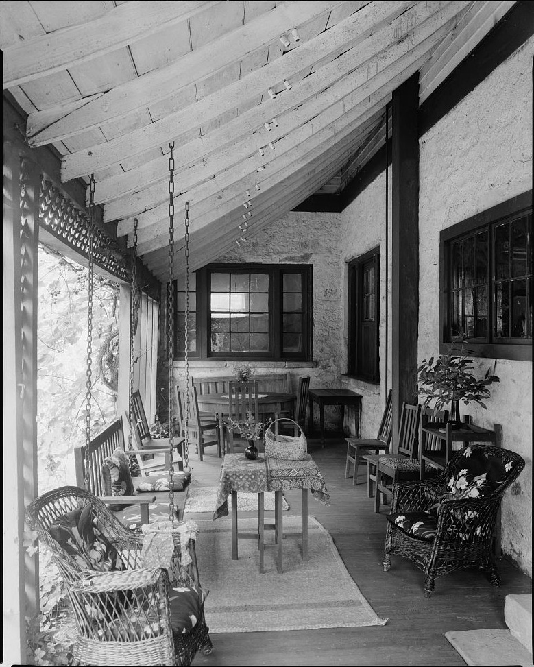 A porch of the Struan house. The walls appear to be stone.  A porch swing faces toward a walk.  Wooden and wicker chairs are along the edges of the porch. A rug laid in the center has a table standing on it. A circular table surrounded by chairs is at one end of the porch. 