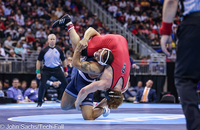 A wrestler maneuvers another wrestler overhead while on a floor mat. Two referees supervise.