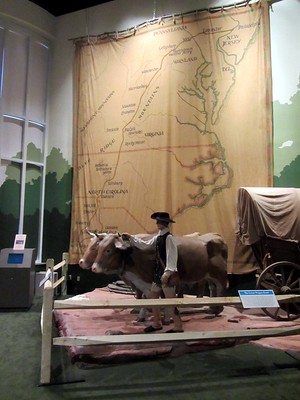 Museum display of a wagon being pulled by two large horses. A model standing next to them. In the background is a large map of North Carolina. 