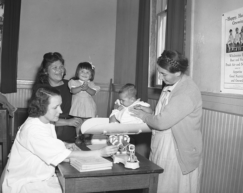 Baby Contest, Cherokee Reservation. Dr. Mary B. Michal examines Lucinda Jane Bradley, 12 Mo old, Nancy Ann Chiltoskie (4 mo old). Weighed by Mrs. Fred Hicks, Public Health Nurse Anderson, Watty, 7 Mos old, Wm. Davis Junaliski, 9 mo old. This photo is part of a collection of photos promoting traveling and tourism in North Carolina, taken by photographers who worked for the Department of Conservation and Development, Division of Travel Information.