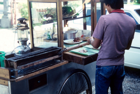 Young man prepares skewered meat for sale from lunch cart