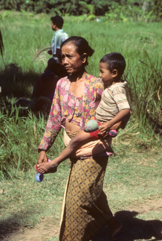 Woman in sarong walks carrying boy on her hip