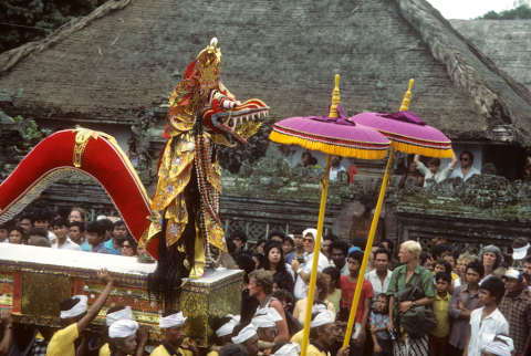 Spectators watch funeral procession of serpent figure and royal umbrellas