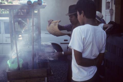 A food stand vendor fans cooking fire as young man watches