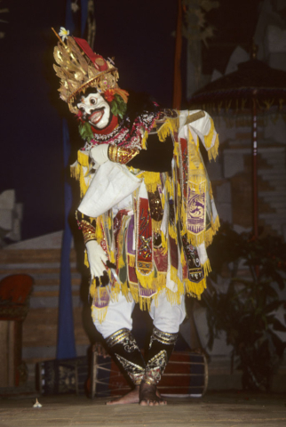 Male masked dancer bows forward slightly and lifts front of tunic