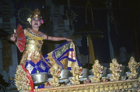 Female Balinese dancer holding open red fan moves behind kettle gong instrument