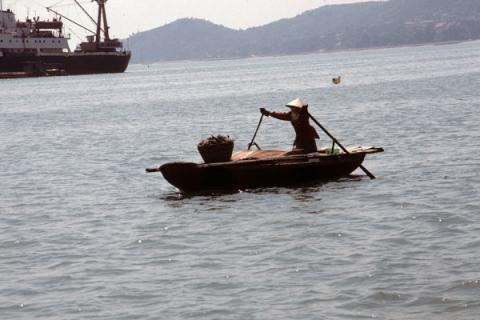 A woman rows a small boat at a distance from two large ships in Halong Bay