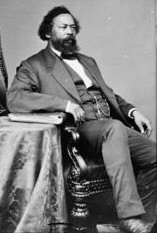 Black and white photograph of a black man with a beard wearing a suit and tie, sitting in a chair. His suit is dark in color and his shirt is white. A pocketwatch chain can be seen at his midsection. 