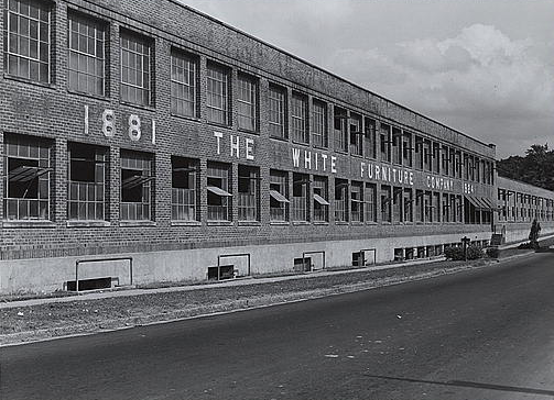 A large warehouse with the name "White Furniture Company" written across the front. It as several large windows and an empty parking lot