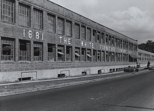 A large warehouse with the name &quot;White Furniture Company&quot; written across the front. It as several large windows and an empty parking lot 